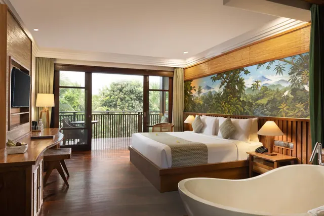 A bedroom with a king bed in front of a painted jungle mural, a freestanding bathtub in the foreground, and glass doors opening onto a balcony with trees beyond.