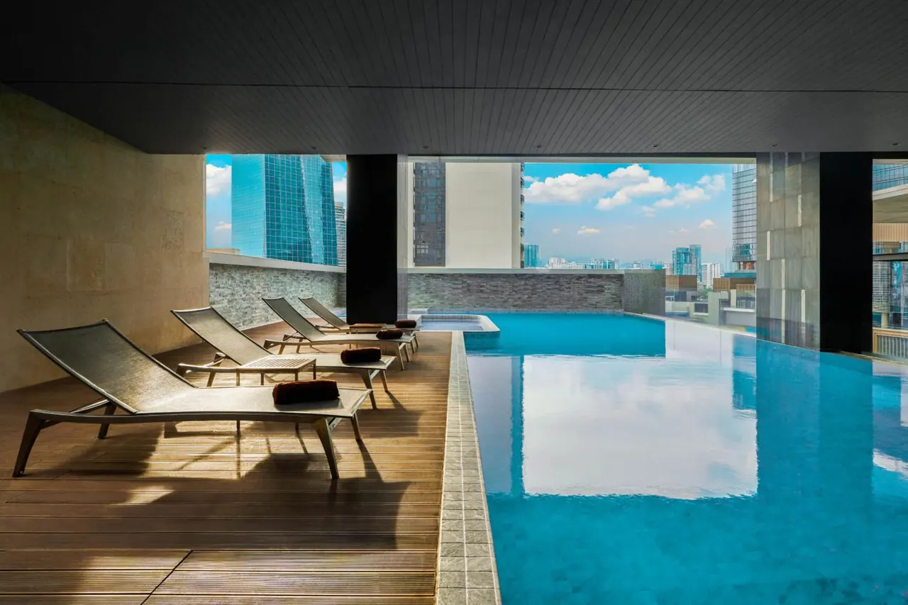 A covered rooftop pool with teal water and timber sun loungers, with city high-rise buildings visible beyond the open side.