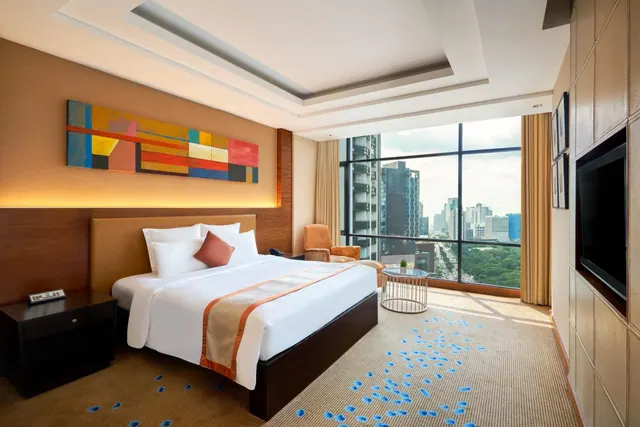 A hotel bedroom with a king bed, a large abstract painting, a wall-mounted television, an armchair, and floor-to-ceiling windows overlooking a city skyline.