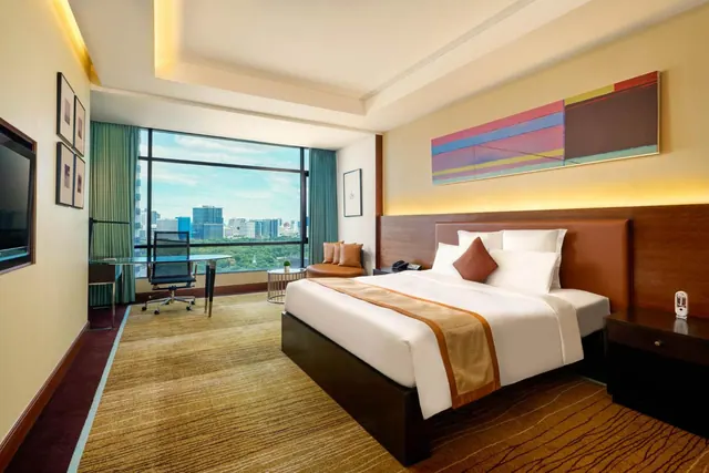 A bedroom with a king bed, a work desk and a seating area by a large window looking out over city buildings and trees.