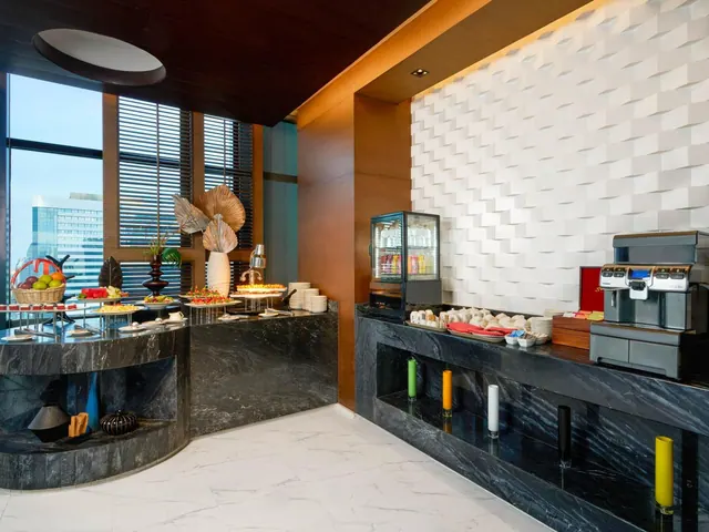 A buffet station with marble counters, food displays, and a coffee machine sits beside a window that looks out onto a glass office building.