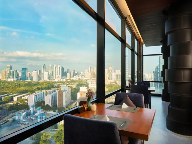 A restaurant dining area with floor-to-ceiling windows overlooking a city skyline, with set wooden tables and dark upholstered chairs.