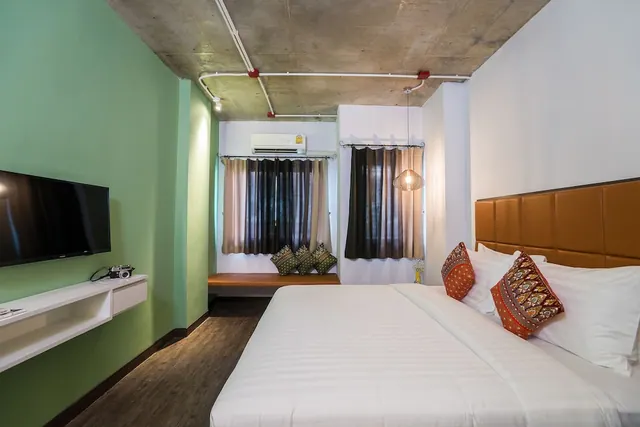 A bedroom with a double bed, a wall-mounted television, a wall air conditioner, and a window seat with cushions under an exposed concrete ceiling.