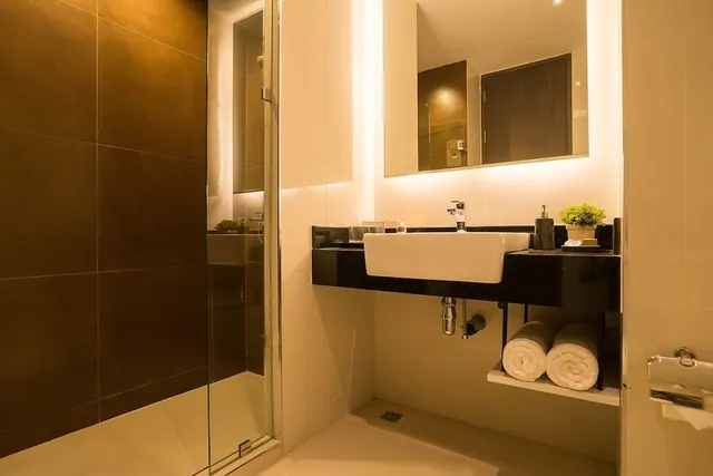 A bathroom with a glass-enclosed walk-in shower, a white vessel sink on a black wall-mounted counter, a backlit mirror and rolled white towels on a shelf.