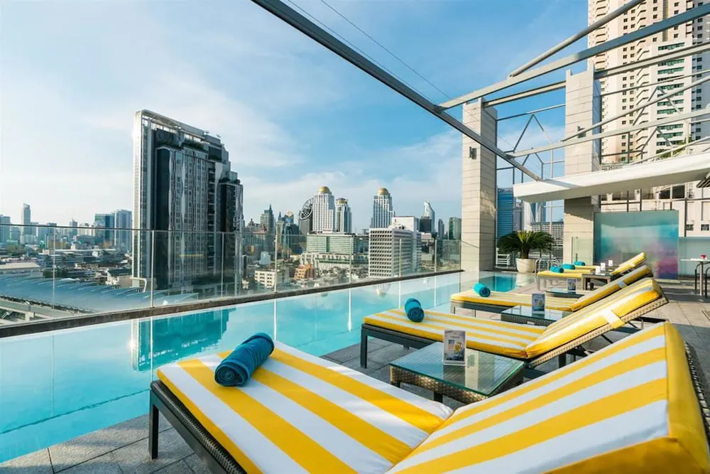 A rooftop infinity pool with yellow striped sun loungers and rolled blue towels overlooks a city skyline of high-rise towers.