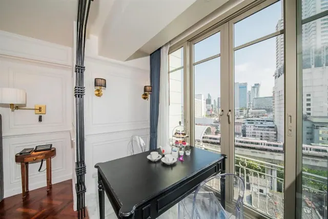 A black dining table set with teacups and an afternoon tea stand sits in a room beside floor-to-ceiling windows that look out over city towers and an elevated train line.