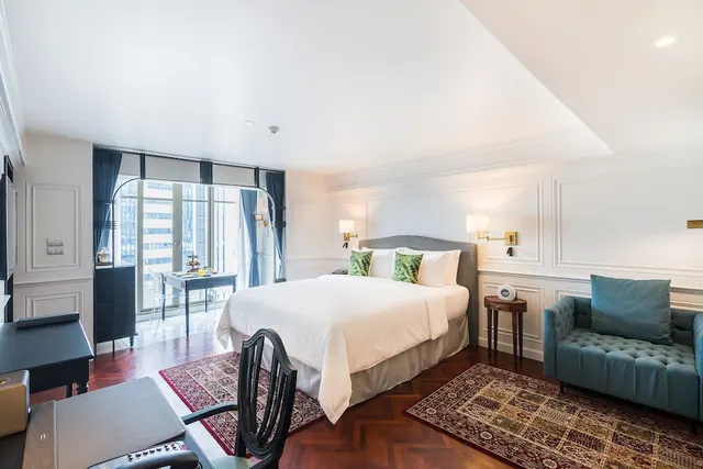 A bedroom with a made double bed, a teal tufted armchair, a desk and glass doors opening onto a balcony with a table and chairs.