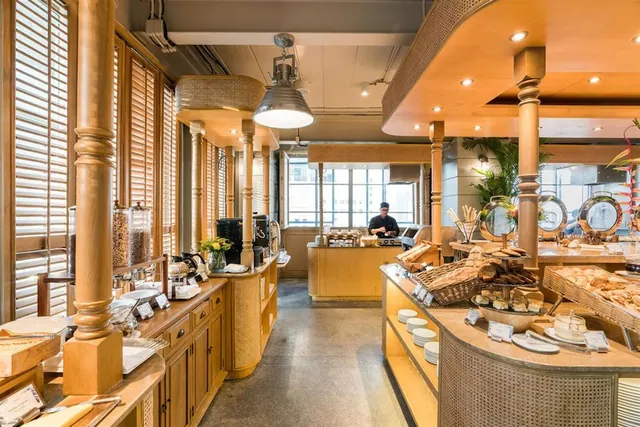 A breakfast buffet restaurant with wooden columns, bread and pastry displays on counters, and a chef working at a station in the background.
