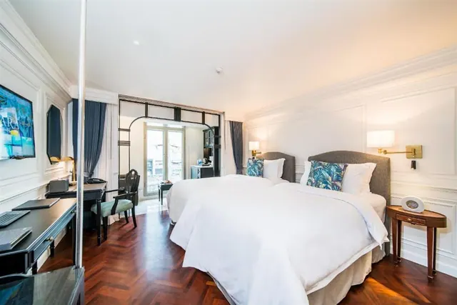 A bedroom with two white-covered beds, a wooden desk with a chair, a wall-mounted television, and glass doors leading out toward a balcony.