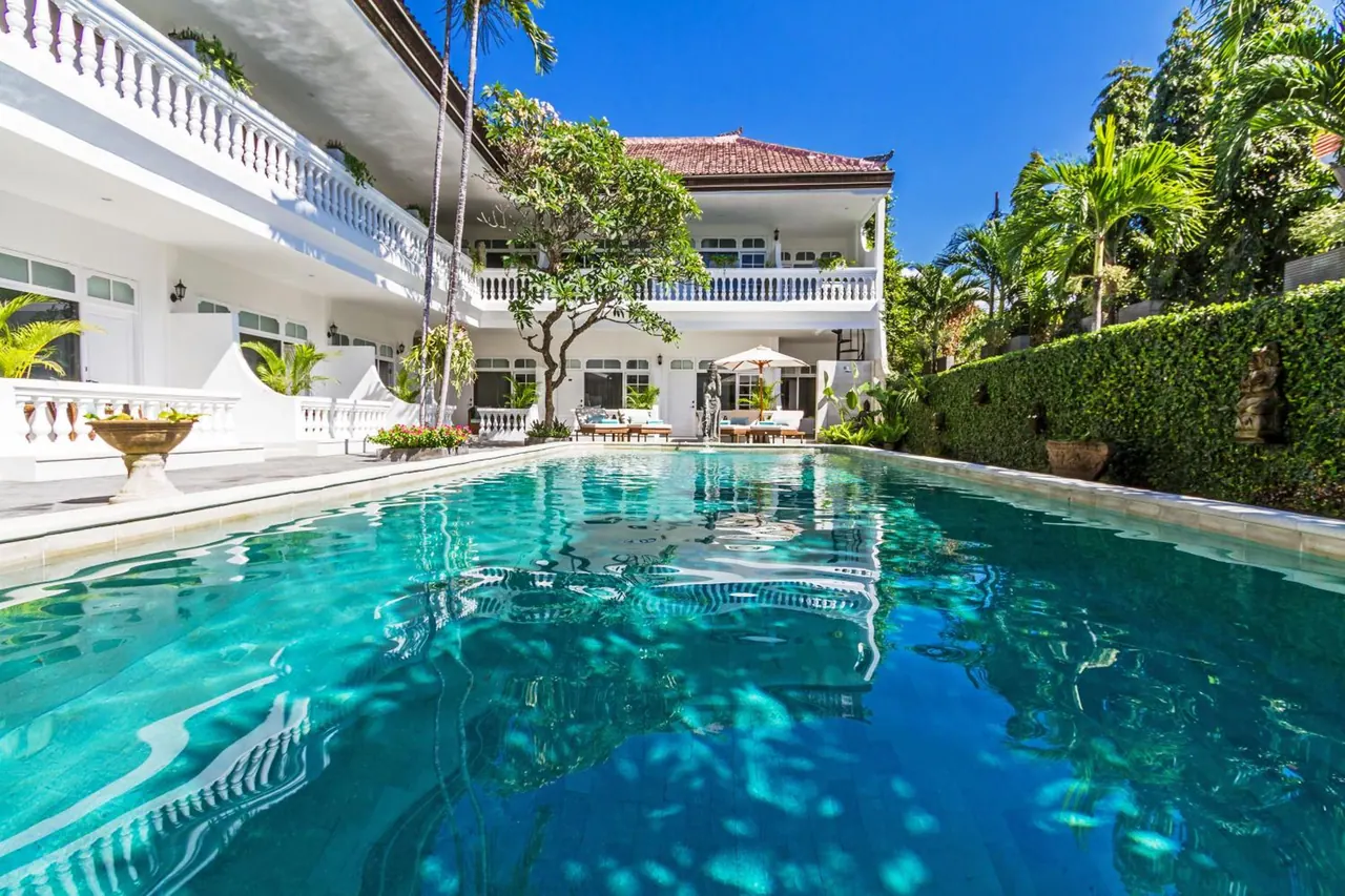A large turquoise swimming pool in front of a white two-storey villa with balconies, sun loungers and a white umbrella under a clear blue sky.