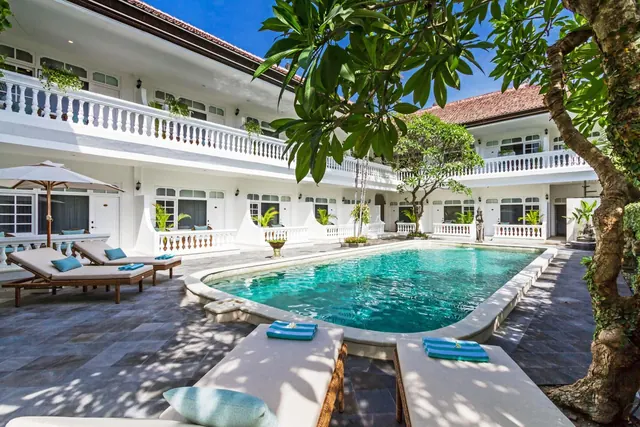 A pool in an open courtyard surrounded by a white two-storey building with balconies, sun loungers and a shaded umbrella.