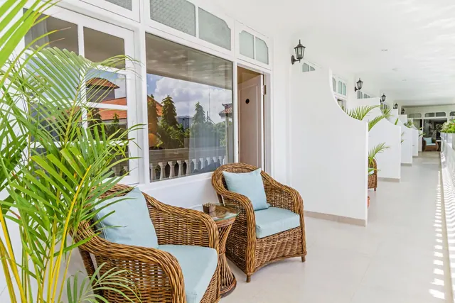 A covered white veranda with wicker armchairs with blue cushions, a glass side table, a potted palm and wall lanterns, with trees and rooftops visible through a window.