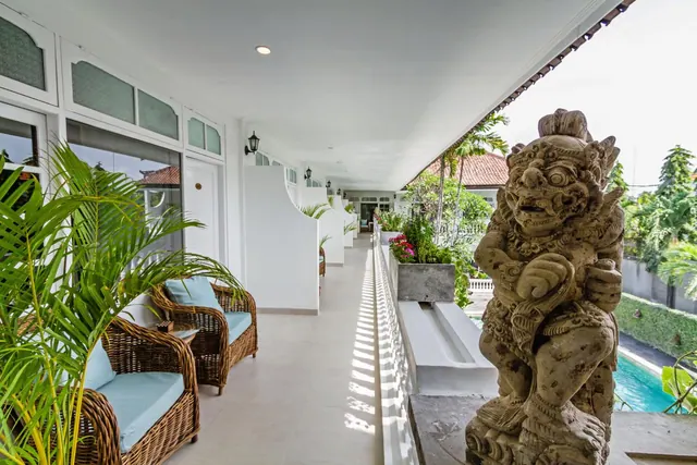 A covered veranda with wicker armchairs and potted palms runs alongside a stone guardian statue, with a swimming pool and trees visible at the right.