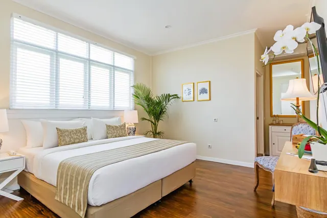 A bedroom with a large bed with white linen, a wooden desk and chair, a wall-mounted television, and a large window with closed white blinds.