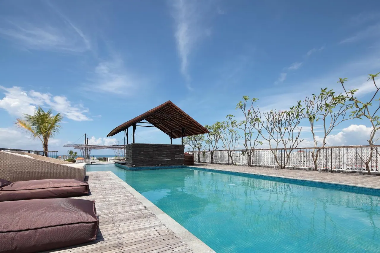 A rooftop lap pool with a wooden deck, cushioned sun loungers, a thatched pool bar pavilion, frangipani trees and a white railing under a blue sky, with the sea visible in the distance.