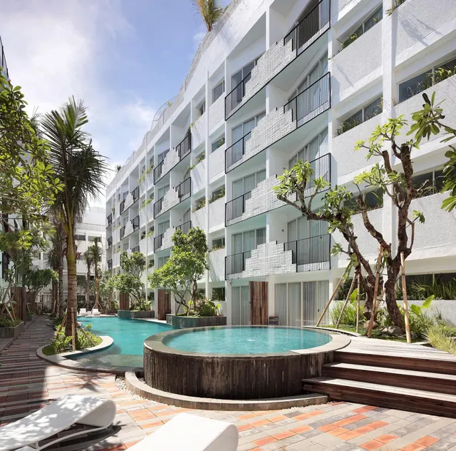 A round sunken pool in the foreground and a longer pool to the left sit beside sun loungers, palm trees and a white multi-storey building with balconies.