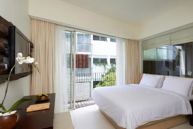 A white king bed sits in a bedroom beside sliding glass doors that open onto a small balcony overlooking a neighbouring building and greenery.