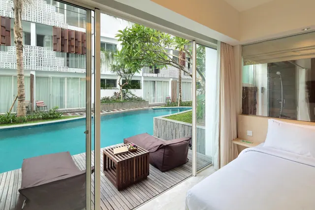 A bedroom with a made bed and sliding glass doors opening onto a timber deck with bean bag seats and a swimming pool beyond, with a glass-walled shower visible at right.