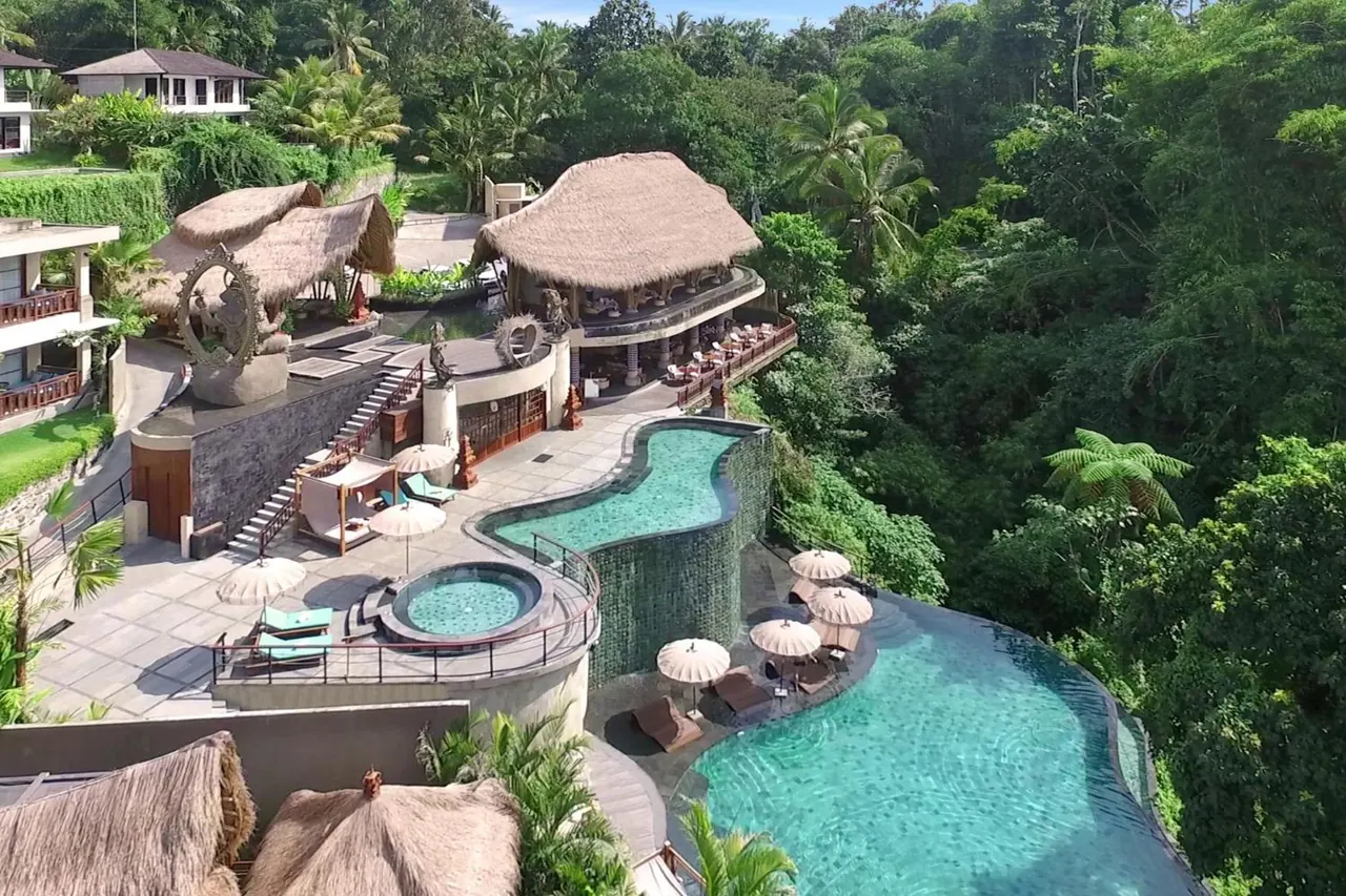 Aerial view of a hotel pool with an infinity edge, sun loungers, umbrellas, a round jacuzzi and thatched-roof buildings surrounded by jungle.