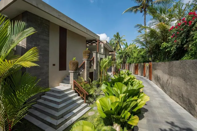 A stone staircase leads up to a modern villa entrance beside a paved walkway, with palm trees and flowering bougainvillea in the background.