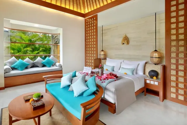 A staged bedroom with a king bed dressed with rose petals, a teal daybed, a round wooden coffee table, and a window seat looking onto green tropical foliage.