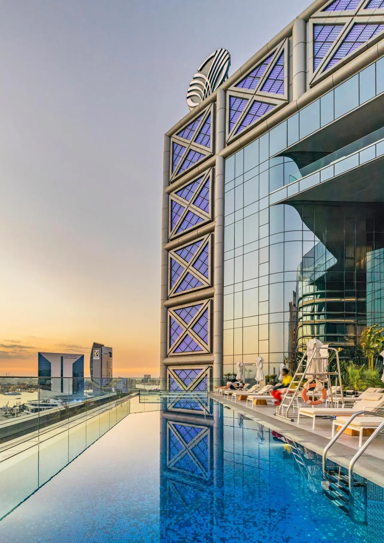 A rooftop infinity pool at dusk runs alongside a tall glass building with geometric window frames, with sun loungers and a city skyline in the background.