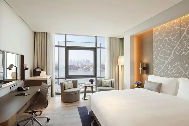 A king bed, work desk and armchairs in a hotel bedroom with floor-to-ceiling windows overlooking water and a distant city skyline.