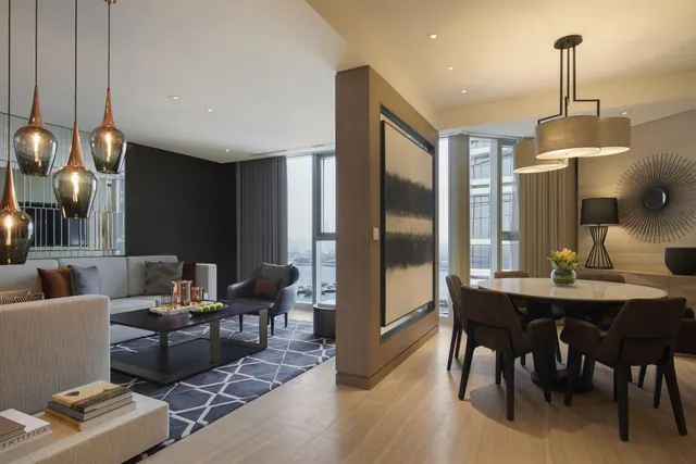 A furnished living area with a sofa, coffee table, round dining table under pendant lights, and large windows showing water and buildings outside.