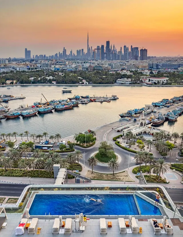 Aerial view from a hotel pool deck over Dubai Creek with dhow boats and the city skyline, including the Burj Khalifa, at sunset.