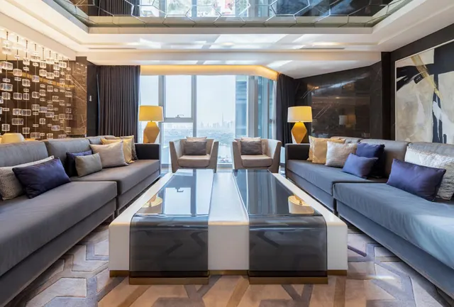 A living room with two grey sofas, a central glass coffee table, two armchairs and floor-to-ceiling windows overlooking a city skyline.