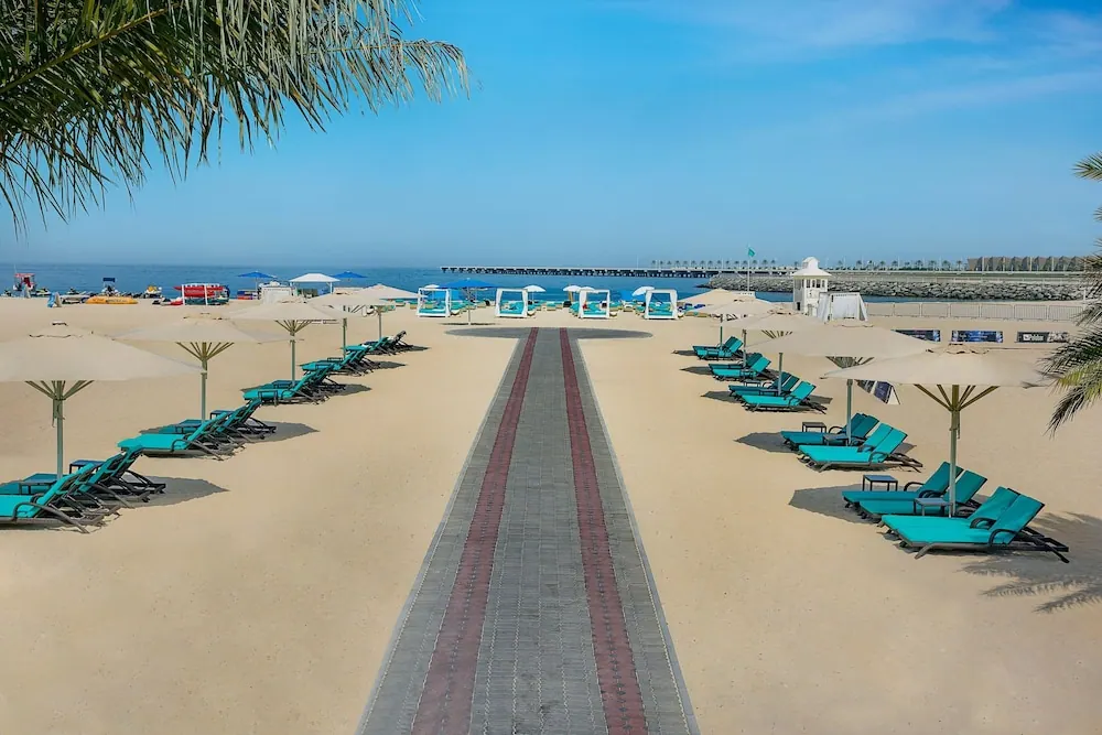 Rows of turquoise sun loungers and cream umbrellas line a paved path that leads down a sandy beach toward the sea and a pier.