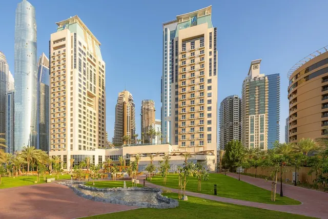 The photo shows a hotel tower and a landscaped park with a fountain, palm trees and grass, surrounded by high-rise towers under a clear blue sky.