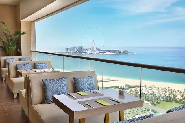 A covered terrace with booth seating and set tables overlooks the sea, a sandy beach, and the Ain Dubai ferris wheel.