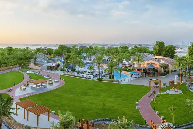 Aerial view of a landscaped resort garden with a lawn, wooden pergolas, a pool area with loungers and umbrellas, a children's play structure, and the sea and city skyline in the background.