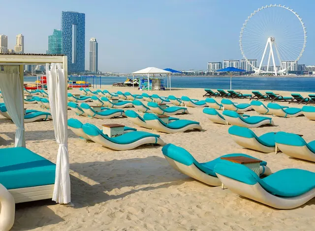 Rows of teal sun loungers and a white cabana stand on a sandy beach, with the sea, a city skyline and the Ain Dubai observation wheel in the background.