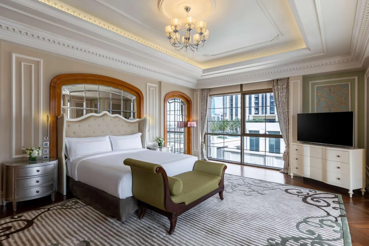 A bedroom with a king bed, a green bench at its foot, a chandelier, a wall-mounted television and windows overlooking high-rise buildings.