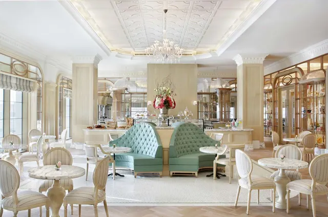 A cream-coloured dining room with a crystal chandelier, green tufted banquettes, marble-top tables with cream chairs, and glass display cabinets along the right wall.