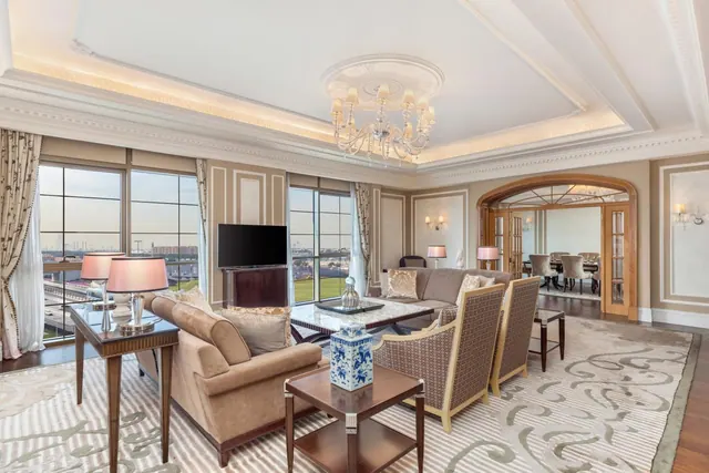 A formal living room with beige sofas and armchairs, a wall-mounted television, a chandelier, and tall windows looking out over distant buildings.