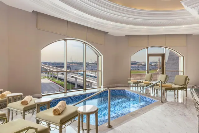 A small indoor pool with blue mosaic tiles sits beside loungers with rolled towels, with arched windows looking out over an elevated metro line and distant city buildings.
