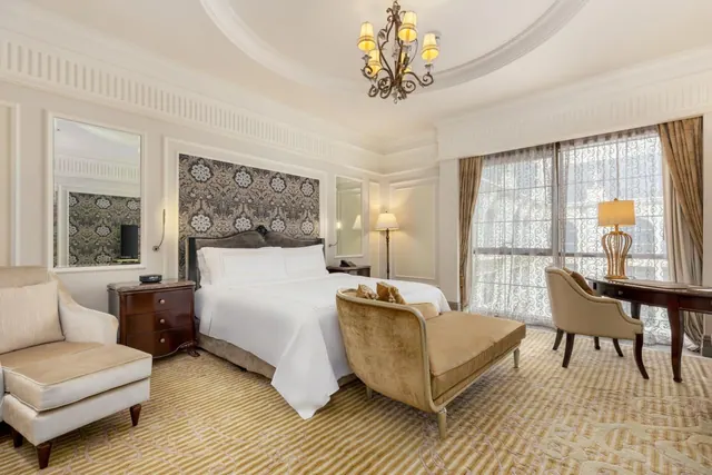A staged bedroom with a king bed, an upholstered bench at the foot of the bed, an armchair, a writing desk by a large window, and a decorative chandelier.