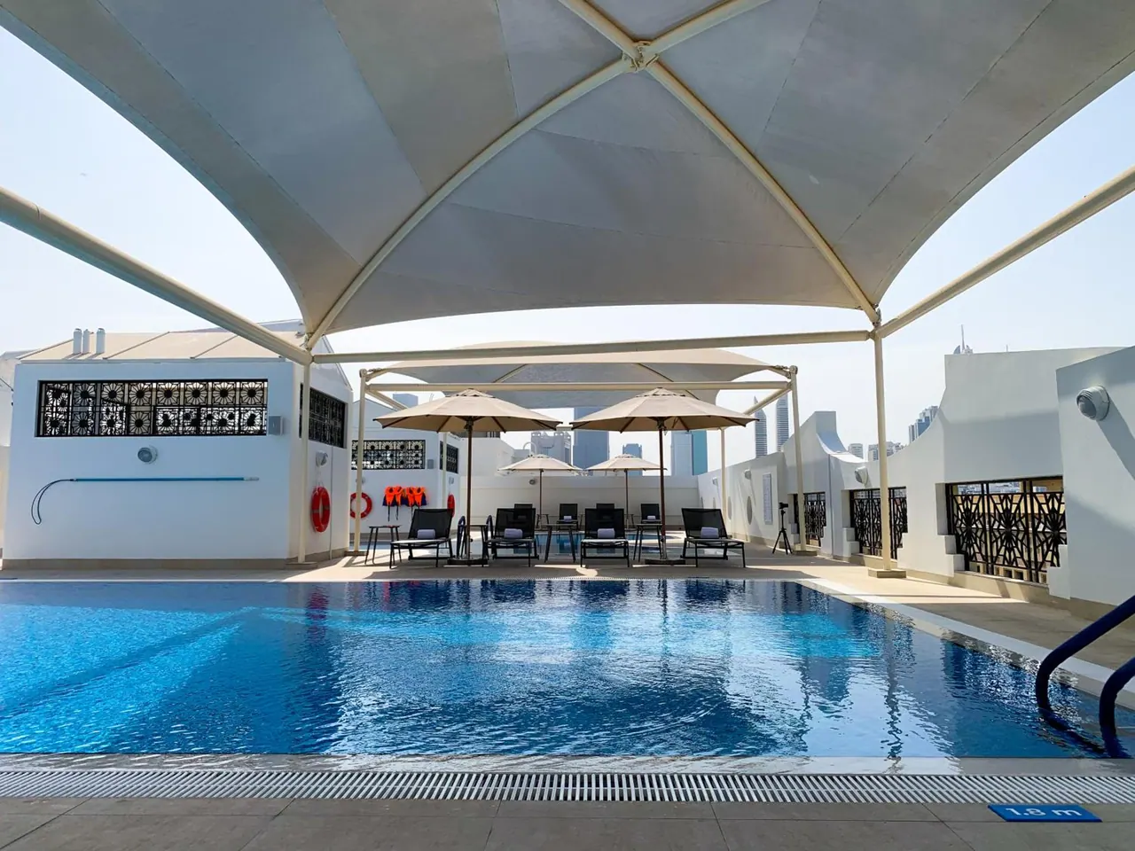 A rooftop swimming pool with lane lines sits under a large fabric canopy, with sun loungers and umbrellas beside it and city towers in the background.