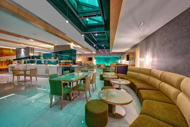 A restaurant dining room with a curved olive-green banquette, green chairs and round tables on a teal marble floor, a buffet counter in the background, and a glass skylight overhead.
