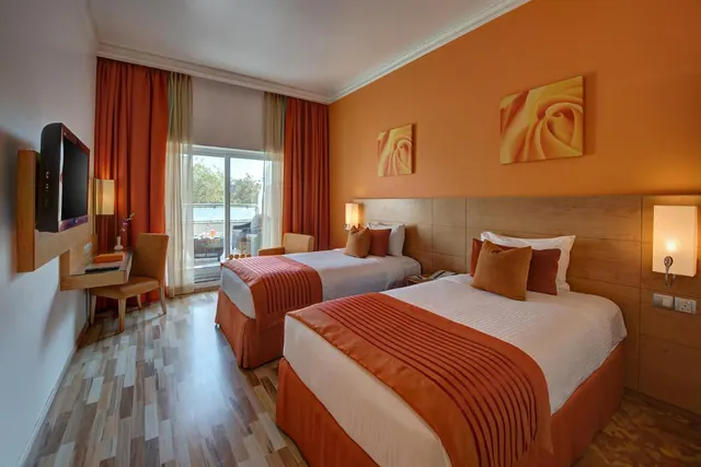 A twin-bedded hotel room with orange walls and curtains, a wall-mounted television, a desk, and a sliding door onto a balcony with trees beyond.