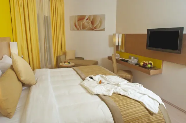 A hotel bedroom with a made bed topped with a white bathrobe, yellow curtains, a small seating area, a wall-mounted desk with a chair, and a flat-screen television.