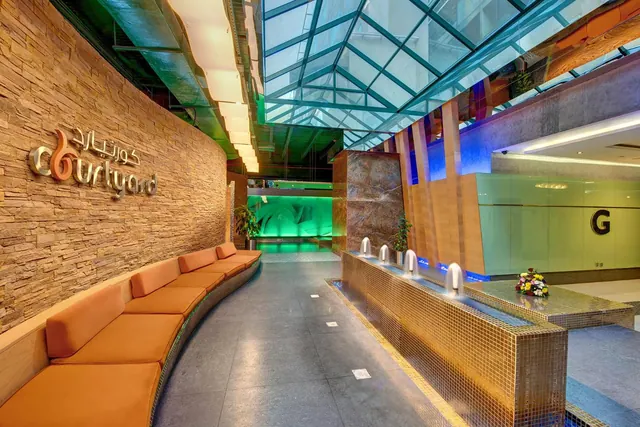 A lobby area with a curved orange bench along a stone wall bearing the Courtyard logo, a glass roof above and a tiled water feature with spouts beside it.