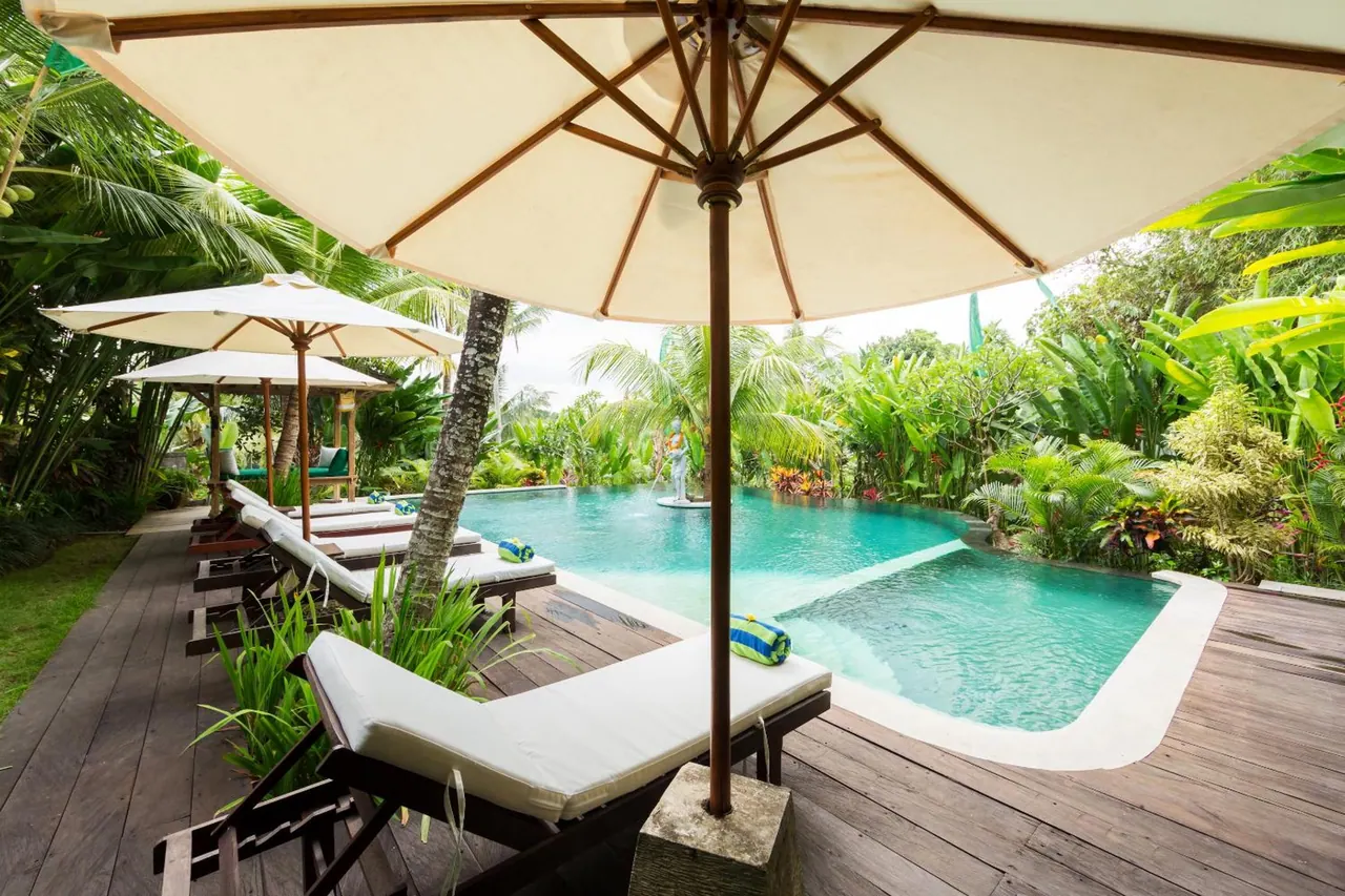 Sun loungers and parasol umbrellas sit on a timber deck beside a turquoise pool, with tropical garden planting behind.