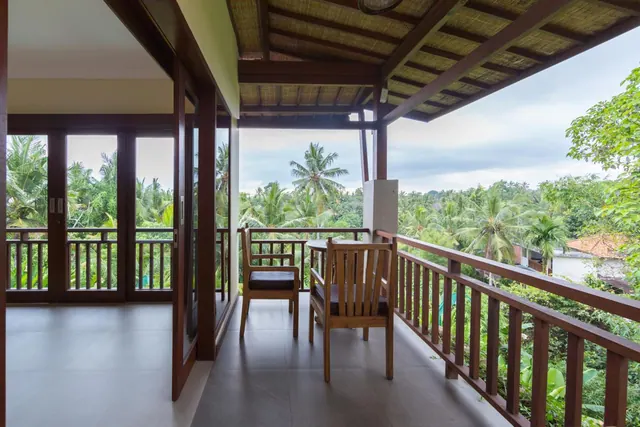 A covered wooden balcony with two chairs and a small table overlooks palm trees and dense greenery, with glass sliding doors into the room on the left.