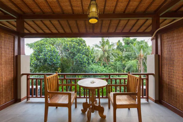A covered balcony with two wooden armchairs, a round wooden table, a hanging woven pendant lamp, and a view of tropical trees and palms beyond the railing.
