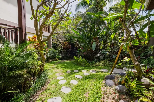 A stepping-stone path crosses a grassy garden lined with tropical plants and a frangipani tree, beside a white building with a wooden railing.