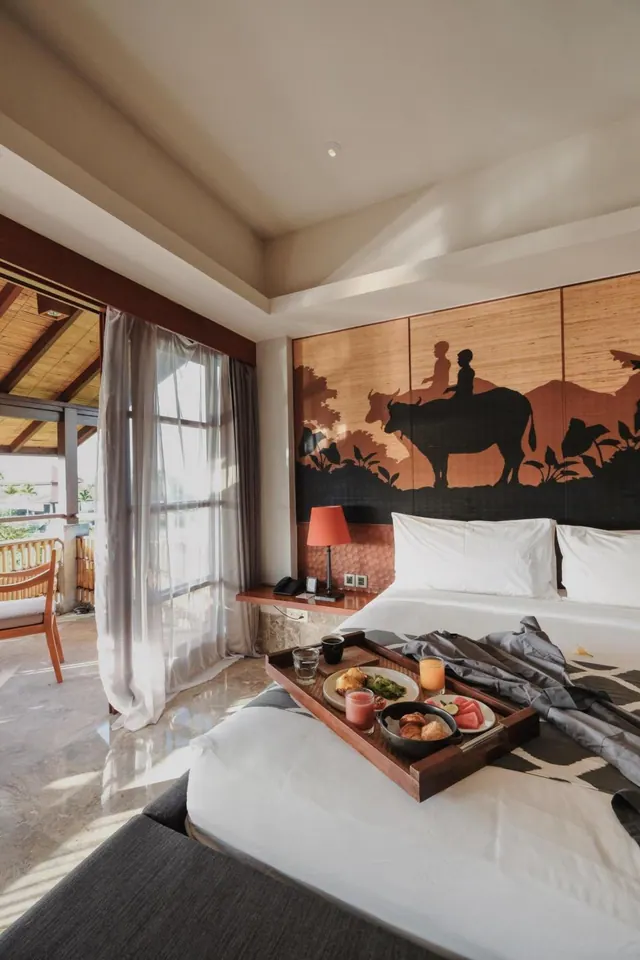 A king bed with a breakfast tray sits in a bedroom with a buffalo silhouette mural on the headboard wall, and sheer curtains are open to a balcony with a wooden chair.